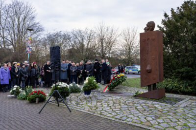 Das Bild zeigt die im Halbkreis aufgestellten Gedenkenden am Mahnmal "MAGDA", vor dem Kränze niedergelegt sind