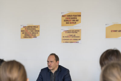 Falko Grube sitzt beim Rollenspiel im Landtag vor eine Gruppe von Schüler*innen, hinter ihm an der Wand kleben gelbe Zettel mit Hinweisen für das Rollenspiel