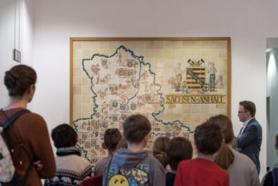 Eine Gruppe von Schüler*innen steht im Magdeburger Landtag vor dem historischen Kachelbild, das eine Karte von Sachsen-Anhalt in seinen alten Grenzen sowie das ehemalige Wappen zeigt