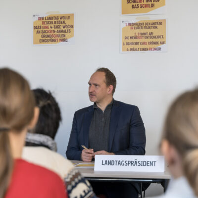 Falko Grube sitzt beim Rollenspiel im Landtag vor eine Gruppe von Schüler*innen, hinter ihm an der Wand kleben gelbe Zettel mit Hinweisen für das Rollenspiel