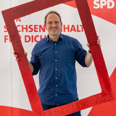 Falko Grube steht lächelnd vor einem Roll-up der SPD Sachsen-Anhalt und blickt durch einen roten Rahmen, den er in den Händen hält.